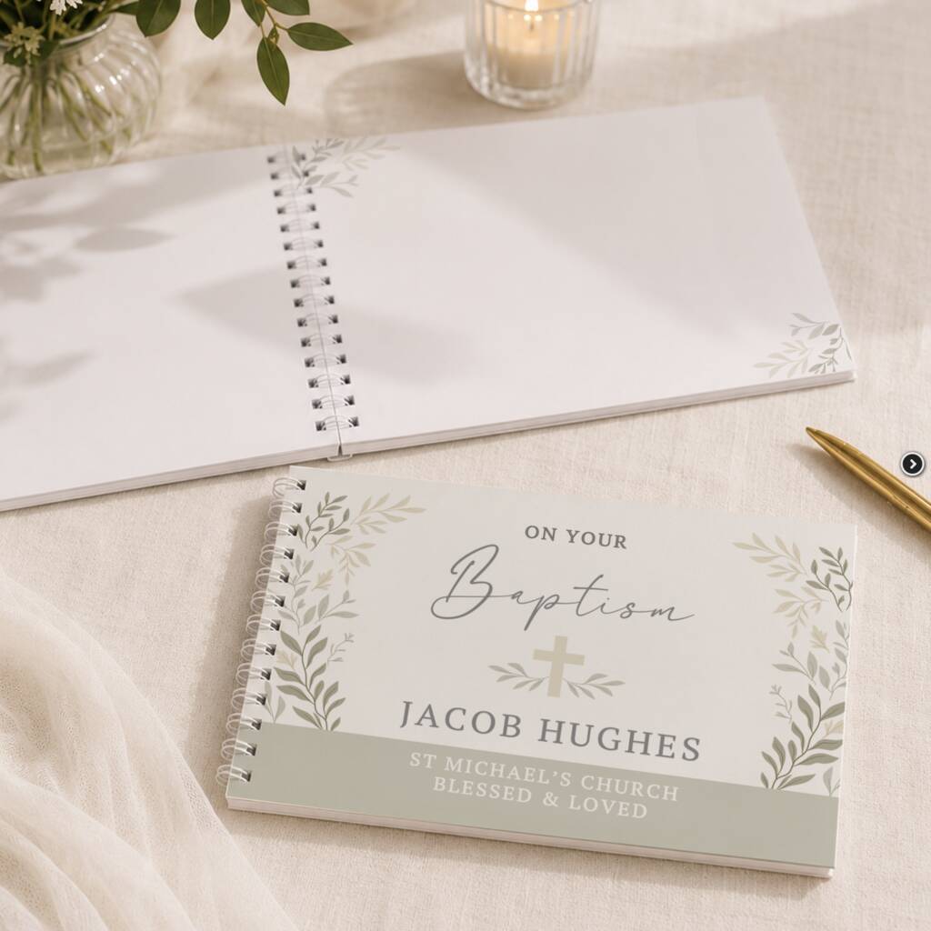 A personalized spiral-bound baptism keepsake notebook lies open on a light fabric surface, with a matching closed notebook in front showing the text 'On Your Baptism Jacob Hughes' and decorative leaf borders. A candle, plant leaves, and a gold pen are softly blurred in the background.