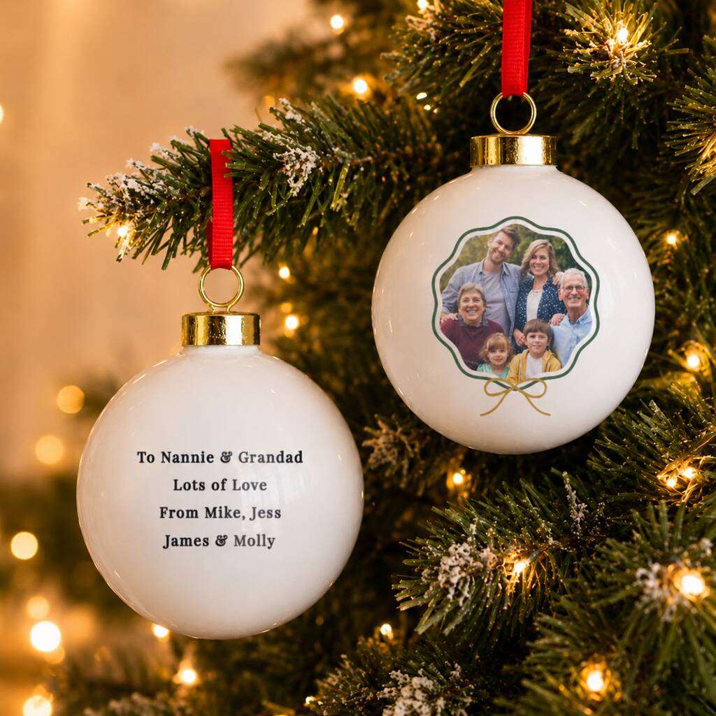 Two personalized white glass Christmas ornaments hanging on a lit Christmas tree. One ornament is printed with the message 'To Nannie & Grandad Lots of Love From Mike, Jess James & Molly' in black text.