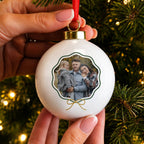 A hand holds a round white personalized Christmas ornament featuring a printed family photo, with a red hanging ribbon and gold cap, set against a blurred Christmas tree background with warm lights.