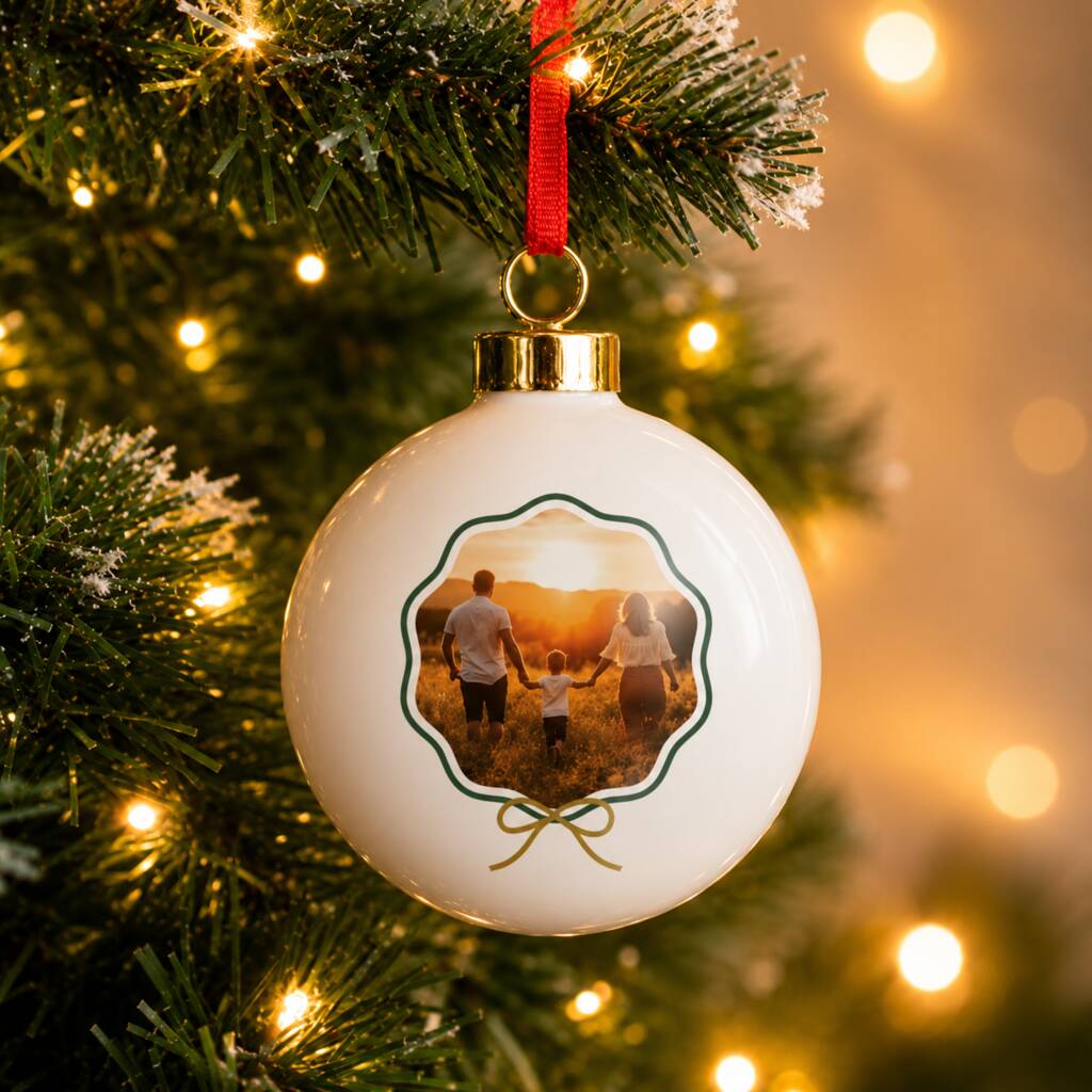 A round white Christmas ornament with a custom family photo printed in the center, hanging from a red ribbon on a decorated Christmas tree with warm lights.