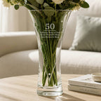 A clear glass vase with a flared shape, engraved with the number 50 and a personalized message, filled with white flowers and green stems, set on a wooden table in a cozy living room setting.