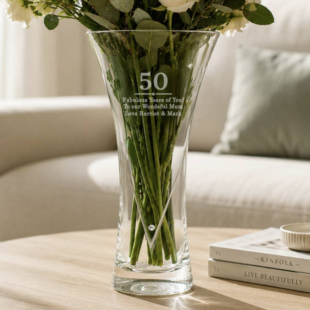 A clear glass vase with a flared shape, engraved with the number 50 and a personalized message, filled with white flowers and green stems, set on a wooden table in a cozy living room setting.