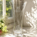 A clear glass vase with a flared top and tapered base, engraved with the number 35 and a memorial message, placed on a windowsill with soft natural light and flowers nearby.