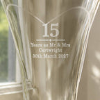 A close-up of a clear glass vase with etched personalization reading '15 Years as Mr & Mrs Cartwright 30th March 2027' in front of a softly blurred window and curtain background.