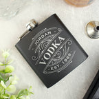 Black hip flask with engraved message on a light surface with plants and glasses.