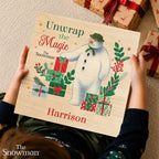 A child holds a personalized wooden book cover featuring an illustration of The Snowman and wrapped gifts, with the text 'Unwrap the Magic', 'The Snowman', and 'Harrison'. The photo is taken from above on a carpet with wrapped presents nearby.
