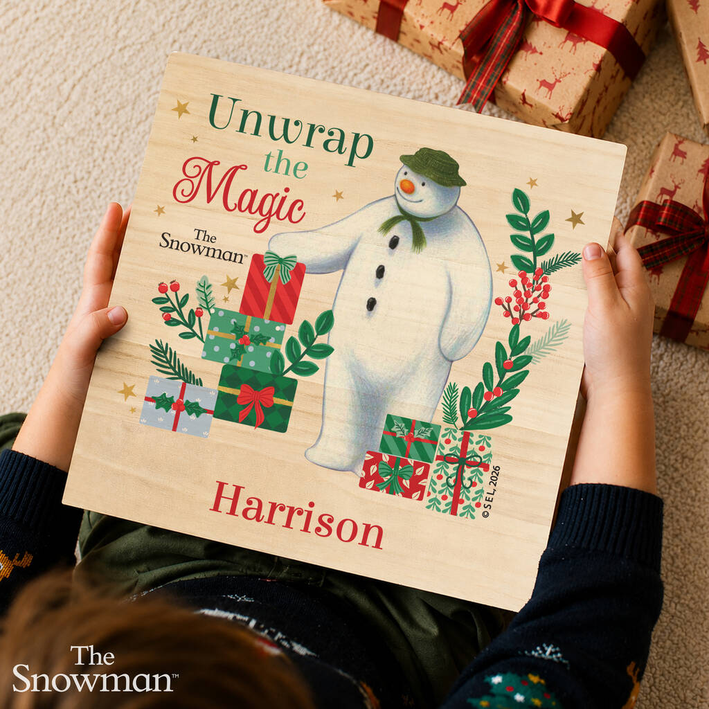 A child holds a personalized wooden book cover featuring an illustration of The Snowman and wrapped gifts, with the text 'Unwrap the Magic', 'The Snowman', and 'Harrison'. The photo is taken from above on a carpet with wrapped presents nearby.