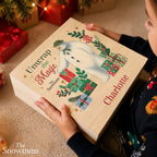A person holds a personalized wooden advent box featuring an illustration of The Snowman and the text 'Unwrap the Magic' and 'Charlotte'. The scene is set in a cozy, festive home environment with Christmas decorations in the background.