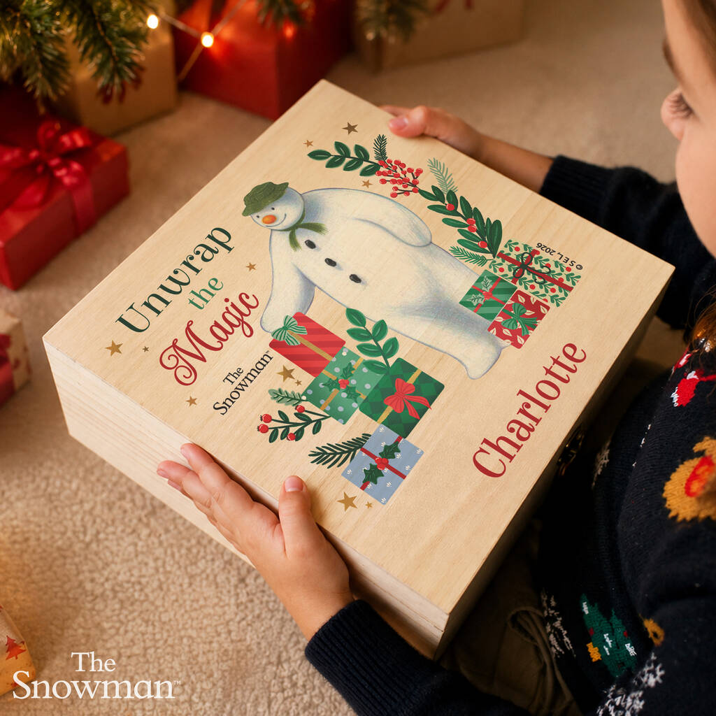 A person holds a personalized wooden advent box featuring an illustration of The Snowman and the text 'Unwrap the Magic' and 'Charlotte'. The scene is set in a cozy, festive home environment with Christmas decorations in the background.