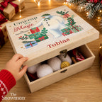 A wooden gift box with a personalized lid reading 'Unwrap the Magic Snowman' and the name 'Tobias', shown open on a wooden table with a child's hand reaching in. The box contains plush white pom-poms and a gold coin, with festive lights and wrapped gifts in the background.
