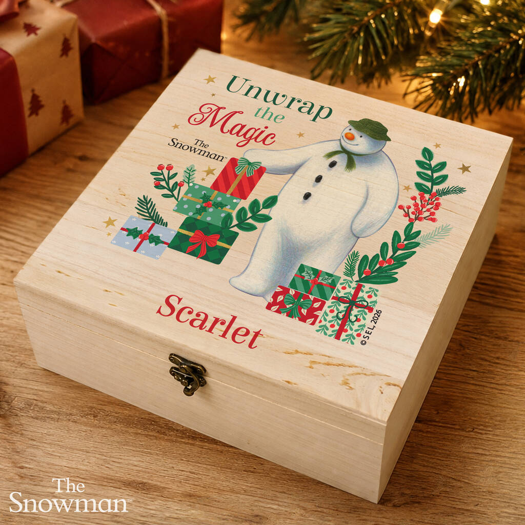 A personalized wooden keepsake box with a Christmas-themed lid design featuring The Snowman, wrapped presents, and the text 'Unwrap the Magic' and 'Scarlet'. The box is styled on a wooden table with holiday decorations in the background.