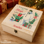 A personalized wooden keepsake box with a Christmas-themed lid design featuring The Snowman, wrapped presents, and the text 'Unwrap the Magic' and 'Scarlet'. The box is styled on a wooden table with holiday decorations in the background.