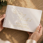 A personalized white spiral-bound wedding guestbook held by two hands over a wooden table. The cover features elegant gold foil script reading 'Wedding Guestbook' and gold printed text with the names 'KYLE & LUCY' and a date.