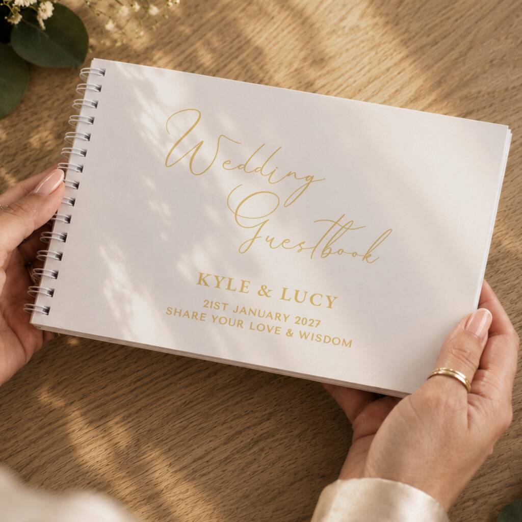 A personalized white spiral-bound wedding guestbook held by two hands over a wooden table. The cover features elegant gold foil script reading 'Wedding Guestbook' and gold printed text with the names 'KYLE & LUCY' and a date.