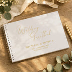 A white spiral-bound wedding guestbook with elegant gold foil lettering, styled on a wooden tabletop with eucalyptus leaves, a cream ribbon, and a gold pen.
