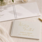 A white spiral-bound wedding guestbook with gold foil lettering reading 'Wedding Guestbook' and names, placed on a light fabric surface next to another open guestbook and a gold pen.
