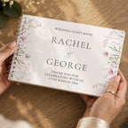 A person holds a spiral-bound wedding guest book with a white cover, elegant black typography, and delicate pink and green floral illustrations along the edges. The book is photographed from above on a warm wooden tabletop with soft natural shadows.