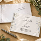 A spiral-bound guest book with a white cover and delicate blue floral illustrations, placed on a wooden table alongside another guest book, a silver pen, and small white flowers.