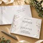 A personalized guest book set displayed on a wooden table with floral accents. The front cover of the spiral-bound guest book reads 'OUR GUEST BOOK SOPHIE & JOANNA' with floral illustrations, and the back cover shows handwritten text.