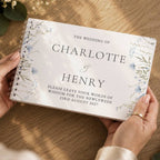 A personalized spiral-bound wedding guest book held by two hands over a wooden table. The white cover features elegant black text reading 'THE WEDDING OF CHARLOTTE & HENRY' with floral illustrations in blue and green along the borders.