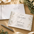 A personalized guest book with floral illustrations on the cover is displayed on a wooden table, alongside another open guest book, a silver pen, and eucalyptus flowers.