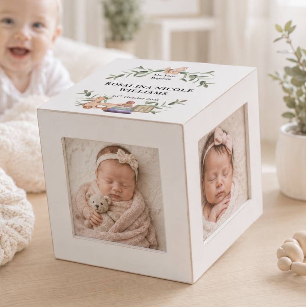 A white wooden cube photo box with three rotating photo frames, shown on a light wood surface with a baby in the background. The top of the box is personalized with the name 'ROMANA NICOLE WELSHANS' and a floral illustration.
