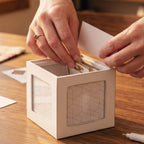 A person's hands placing small gold-toned cards into a white geometric cube organizer with a clear window, set on a wooden tabletop.