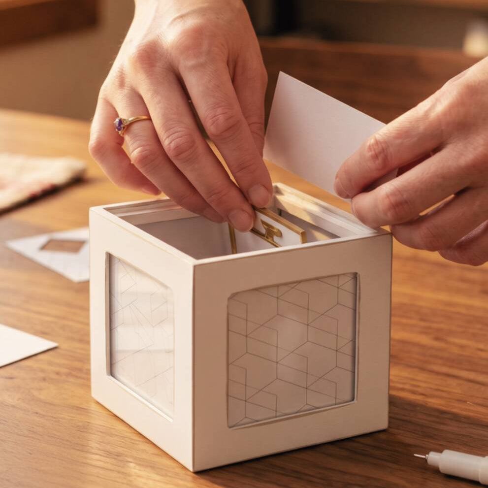 A person's hands placing small gold-toned cards into a white geometric cube organizer with a clear window, set on a wooden tabletop.