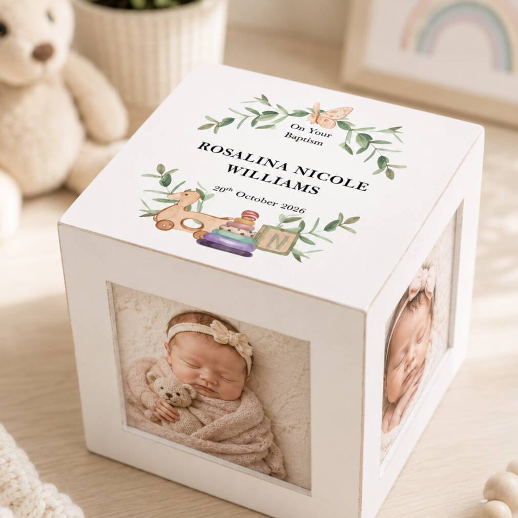 A white square photo cube keepsake box with custom printed photos and text on the lid, styled on a light wooden surface with soft toys and nursery decor in the background.