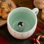 A festive ceramic mug with a small penguin figurine inside, styled on a Christmas table with gingerbread cookies, a candle, and holiday decorations.