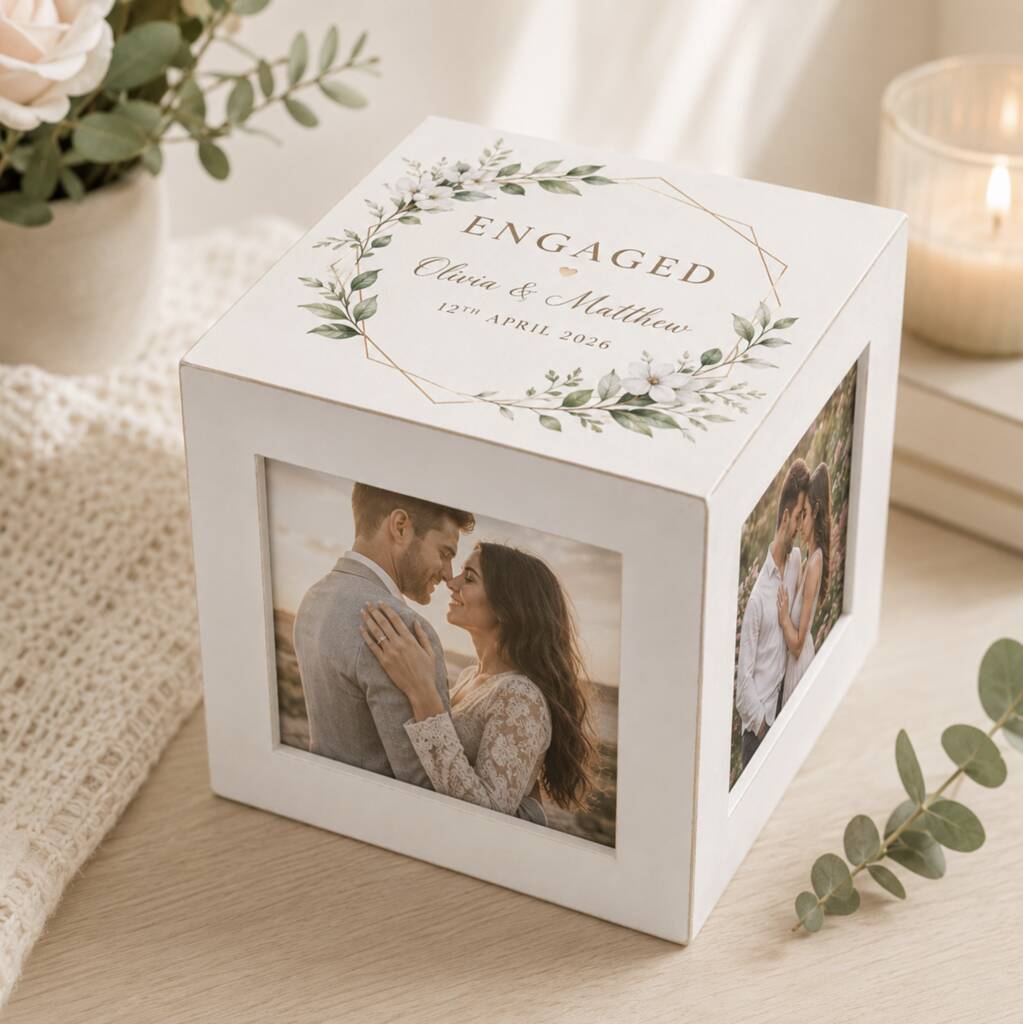 A white personalized engagement photo cube box with floral greenery illustrations and the word 'ENGAGED' on top, shown on a tabletop with a photo of a couple printed on the front and side. The scene includes soft home decor props like a candle, books, and greenery.