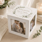A white personalized engagement photo cube box with floral greenery illustrations and the word 'ENGAGED' on top, shown on a tabletop with a photo of a couple printed on the front and side. The scene includes soft home decor props like a candle, books, and greenery.