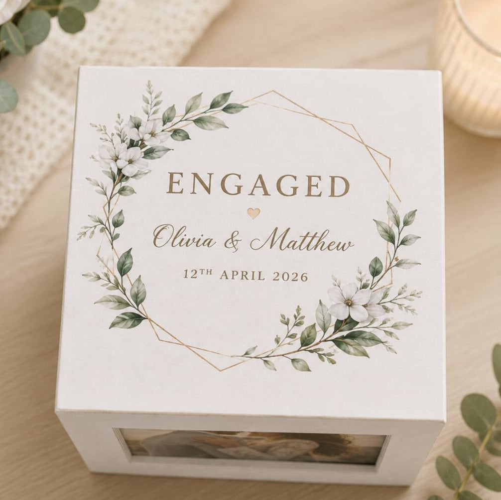 A square white keepsake box with a floral wreath design and the word 'ENGAGED' printed on top, styled on a light wood surface with eucalyptus leaves and a candle in the background.
