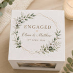 A square white keepsake box with a floral wreath design and the word 'ENGAGED' printed on top, styled on a light wood surface with eucalyptus leaves and a candle in the background.
