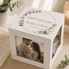 A white square anniversary photo cube box with a personalized top print featuring green floral wreaths and the text 'Anniversary Charlotte & Charles 26th May 2026', shown on a light wooden surface with a photo window on the front displaying a couple and another photo visible on the side.