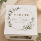 A square white keepsake box with a floral wreath design and the words 'MARRIED', 'Olivia & Matthew', and '12th April 2026' printed on the lid, photographed on a light wood surface with soft decorative props around it.
